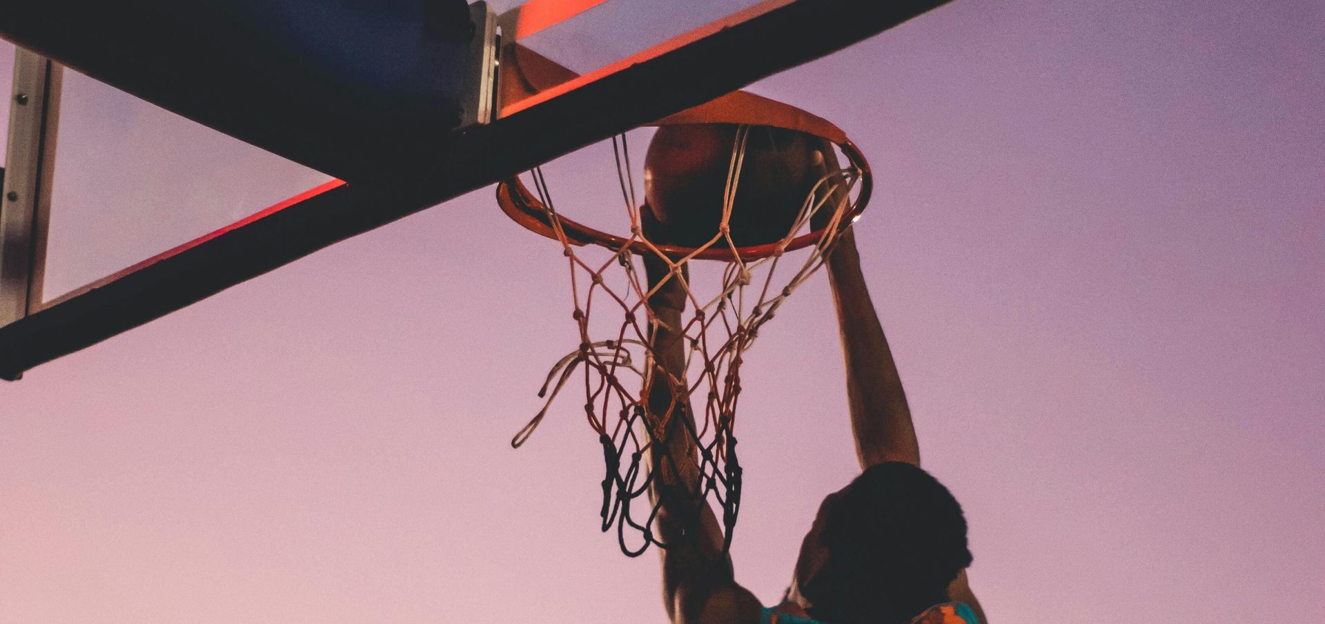 A basketball player successfully dunks as the sun sets, creating a dramatic silhouette.